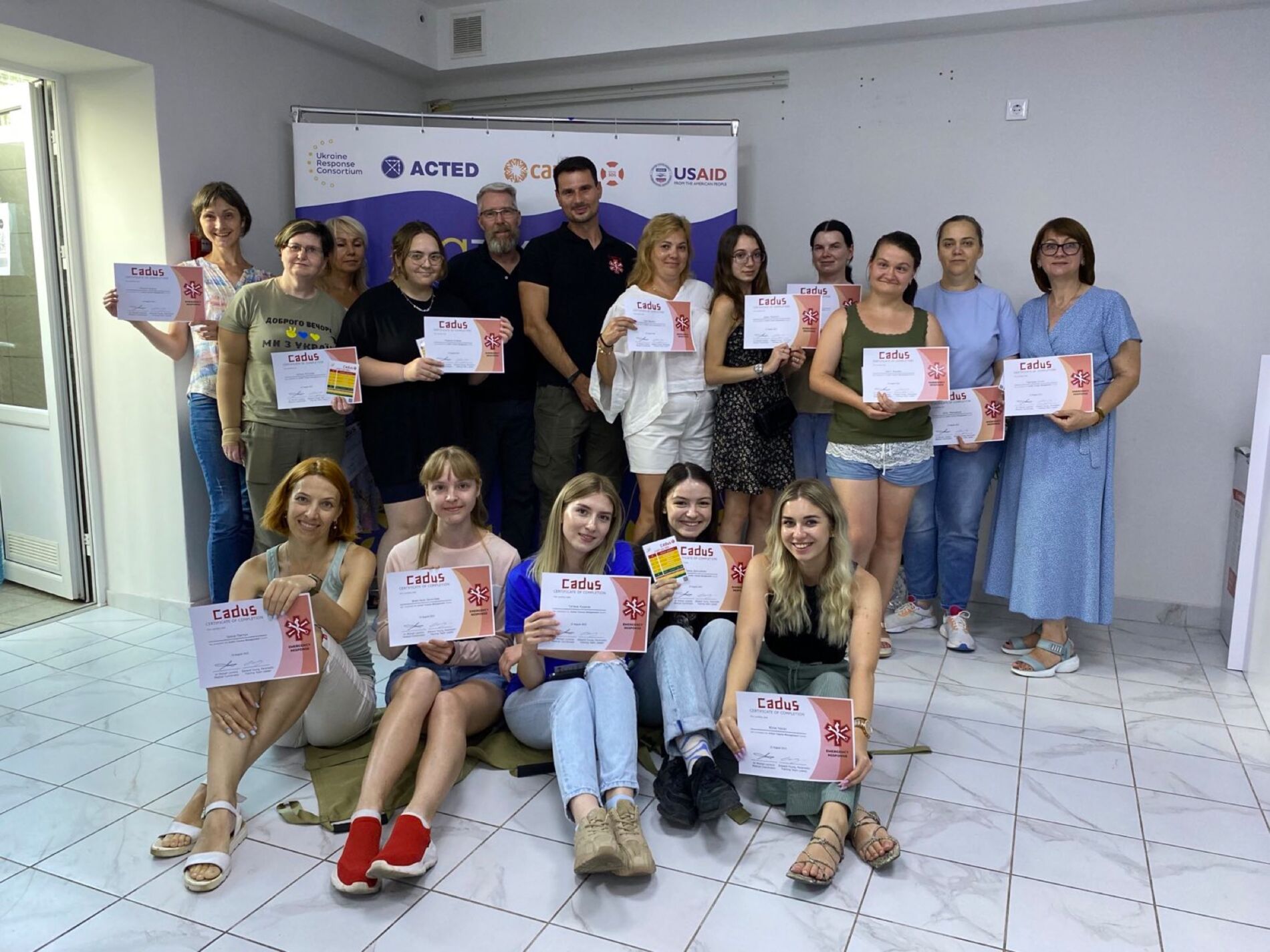 Zwei unserer Trainer inmitten glücklicher Absolventinnen eines Kurses, den sie mit Zertifikat abgeschlossen haben. Foto: CADUS Ein Gruppenfoto in einem Gebäude. In zwei Reihen stehen und sitzen mehrere Absolventinnen eines Kurses, die fröhlich ihre Zertifikate halten. Inmitten der Frauen stehen zwei Kursleiter.