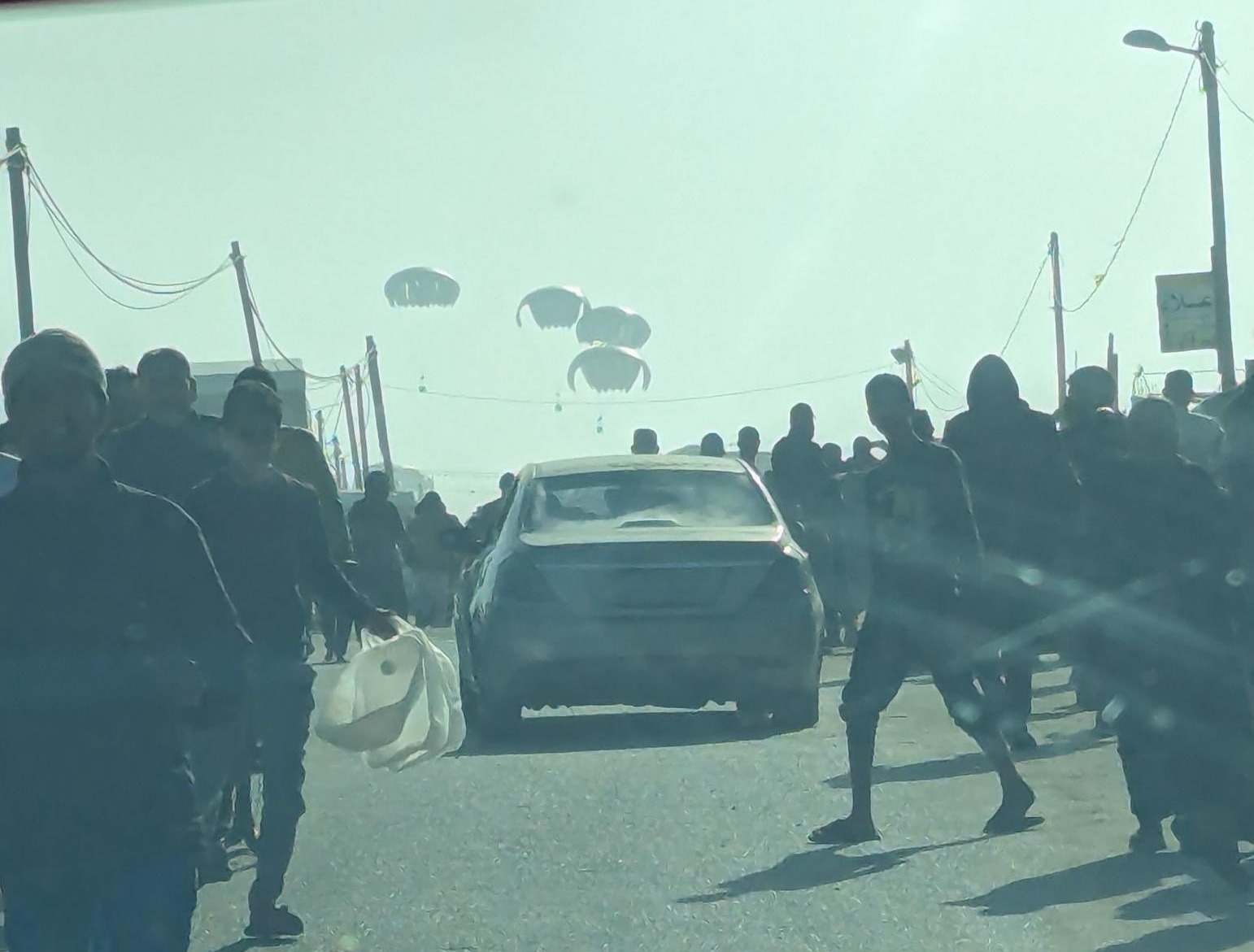Auf einer Straße laufen Menschen links und rechts an einem Auto vorbei in Richtung herabfallender Kisten an Fallschirmen.